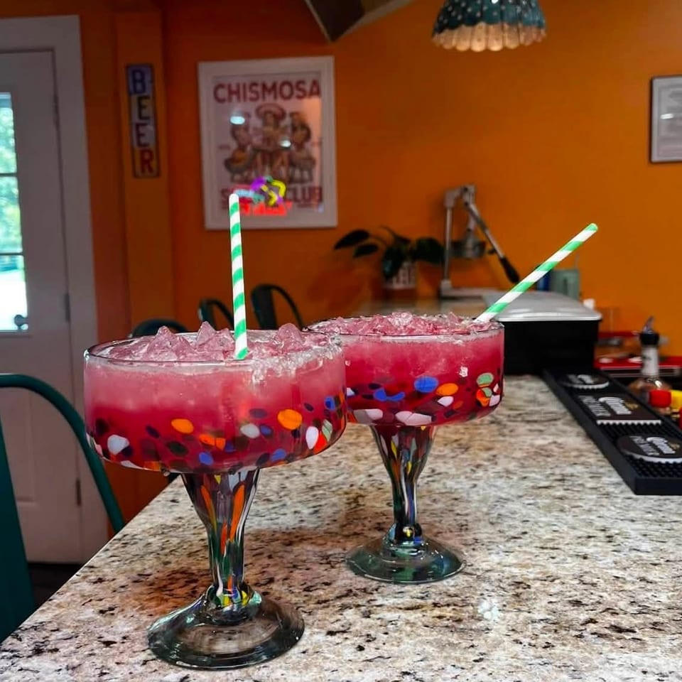 Two bright pink frozen drinks with ice and striped straws sit in colorful margarita glasses on a restaurant bar.