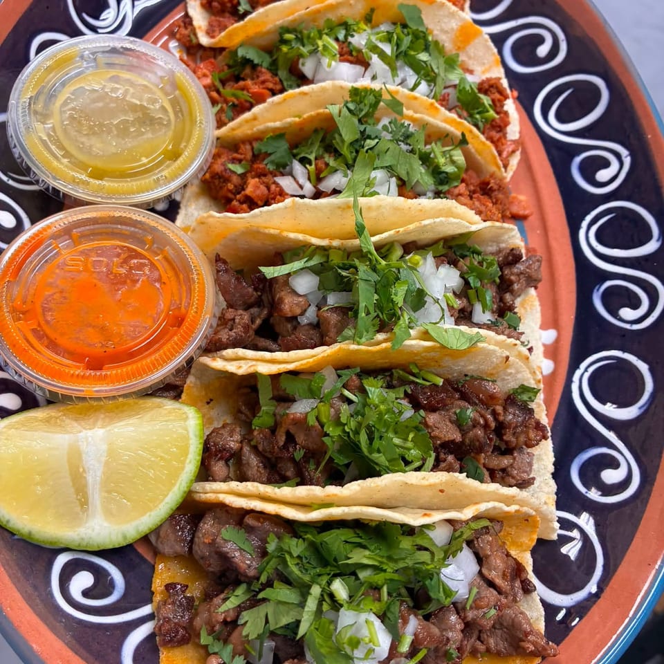 Plate of tacos filled with seasoned meats, onions, cilantro, lime wedges, and cups of red and green salsa.