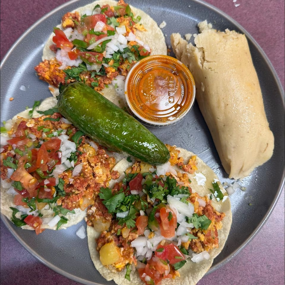 Three tacos topped with seasoned meat, chopped tomato, onion, and cilantro, served with a cucumber, salsa, and a tamale.